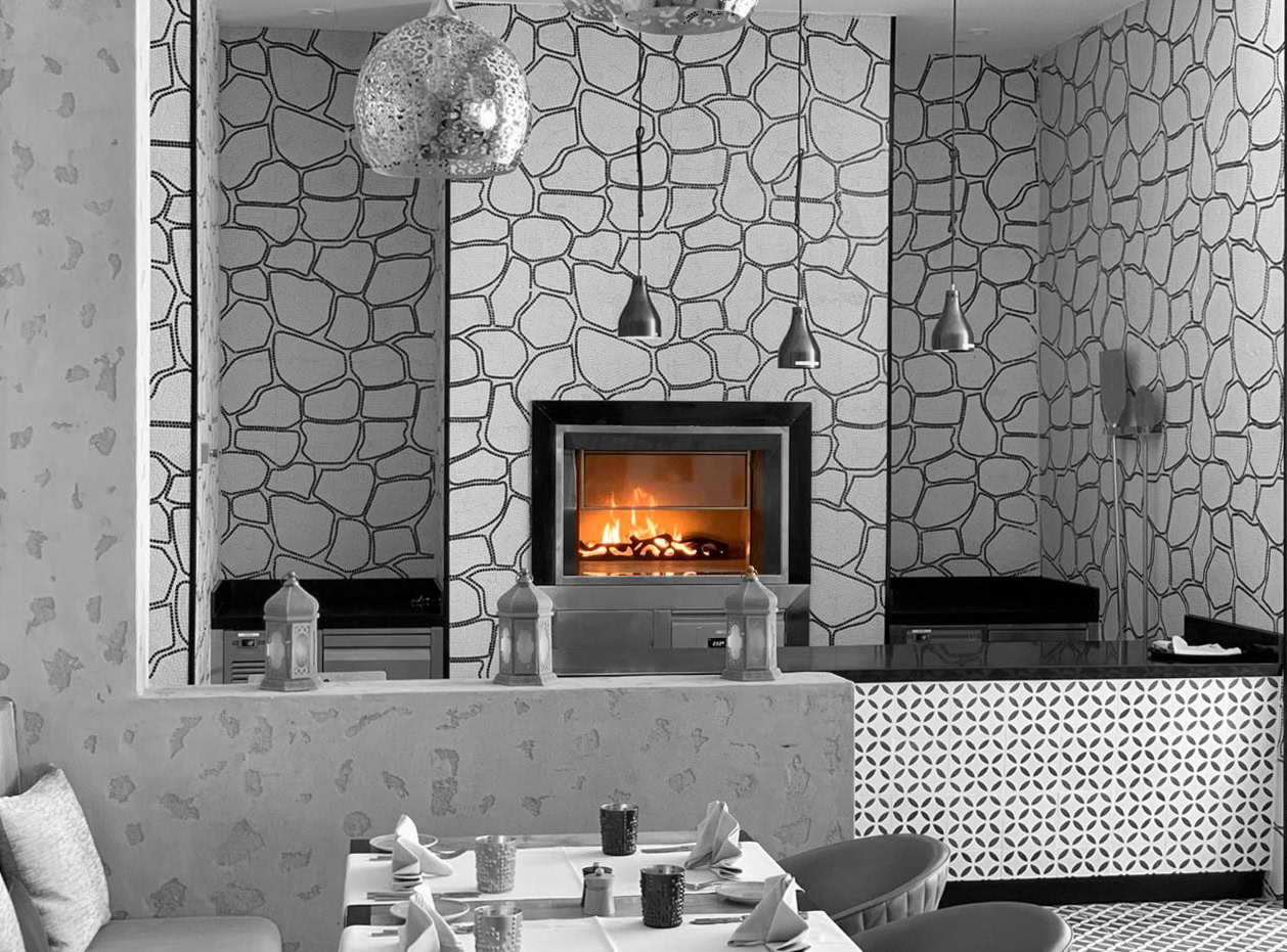 Rectangular stone hearth oven alight in a wall clad in white mosaic stone, under pendant lamps